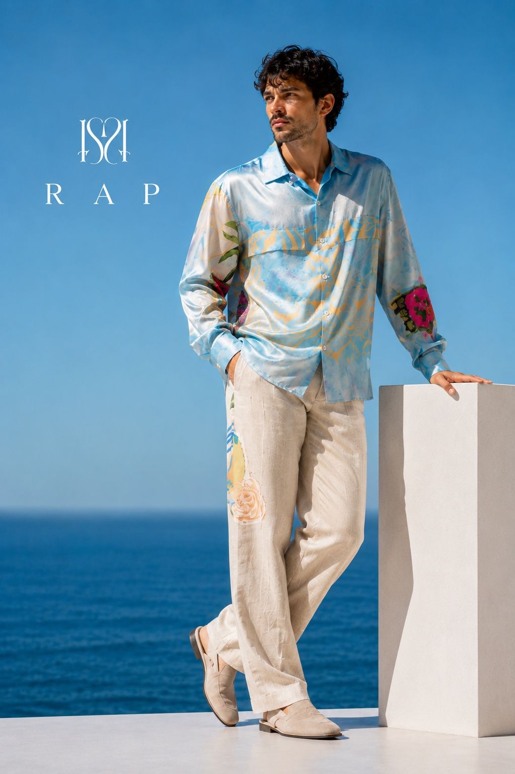 Model wearing a pale blue printed shirt and cream trousers from the Six Season collection beside the sea.