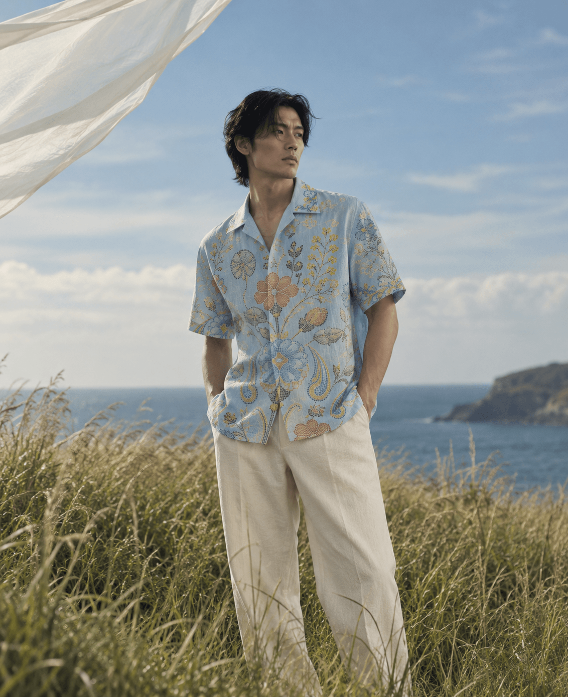 Man wearing a light-blue floral camp-collar shirt and cream trousers in coastal grass.